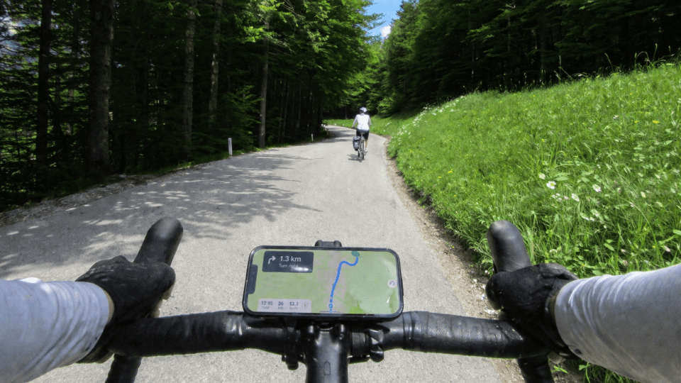 First-person view of a cyclist riding along a winding forest road, with hands on the handlebars and a smartphone mounted in landscape mode displaying navigation, while another rider cycles ahead under dappled sunlight.